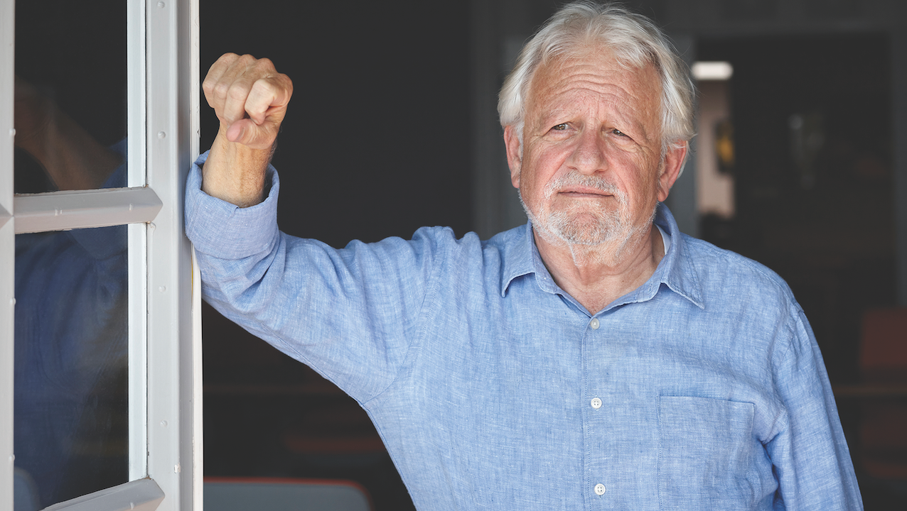 An older man with white hair leaning up against a door looking outside