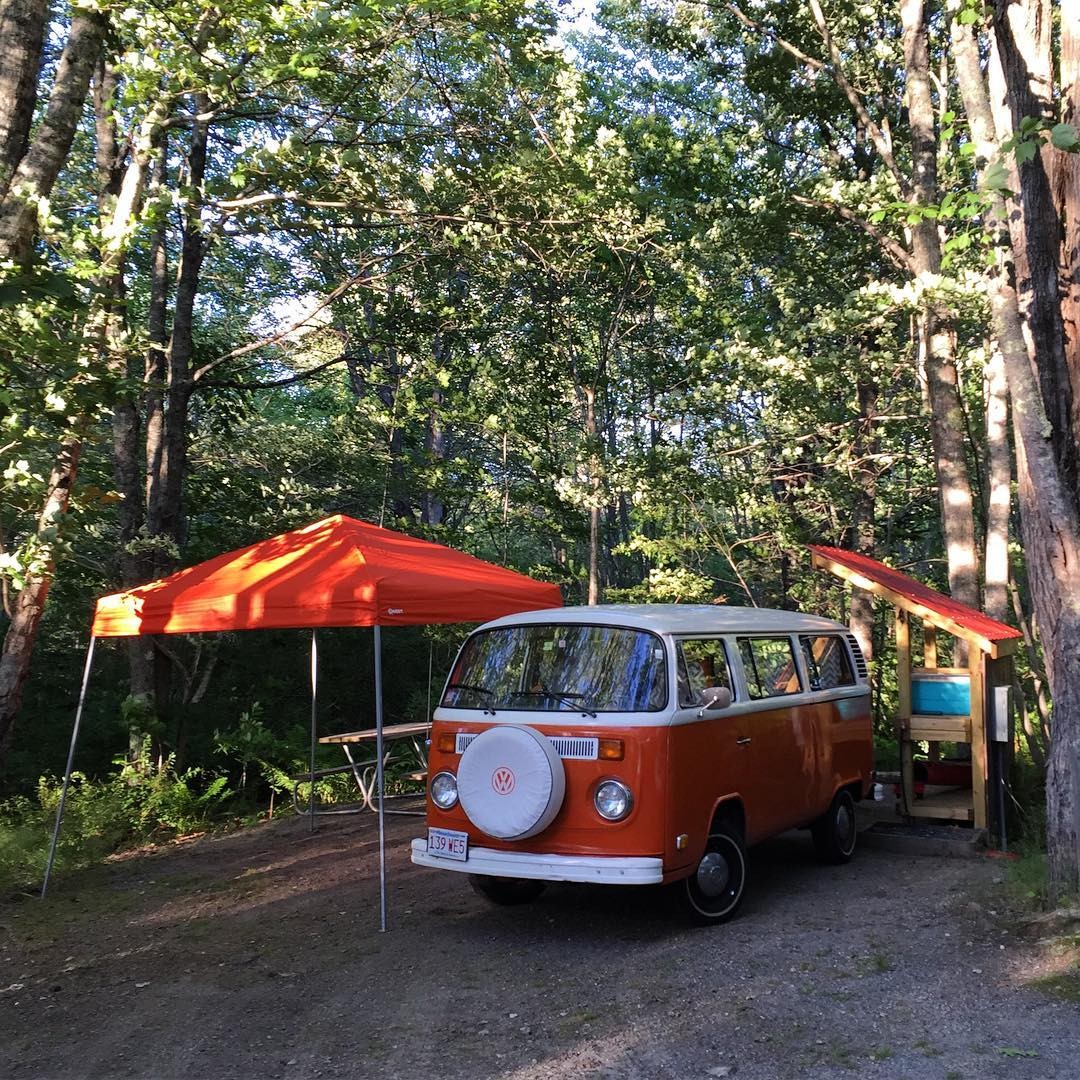 An old orange bus in a forest campground with an orange tent and shelves with an orange roof on each side