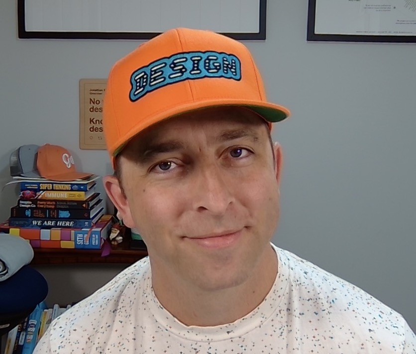 An adult male sitting in a home office wearing a t-shirt a hat with 'design' written across the front