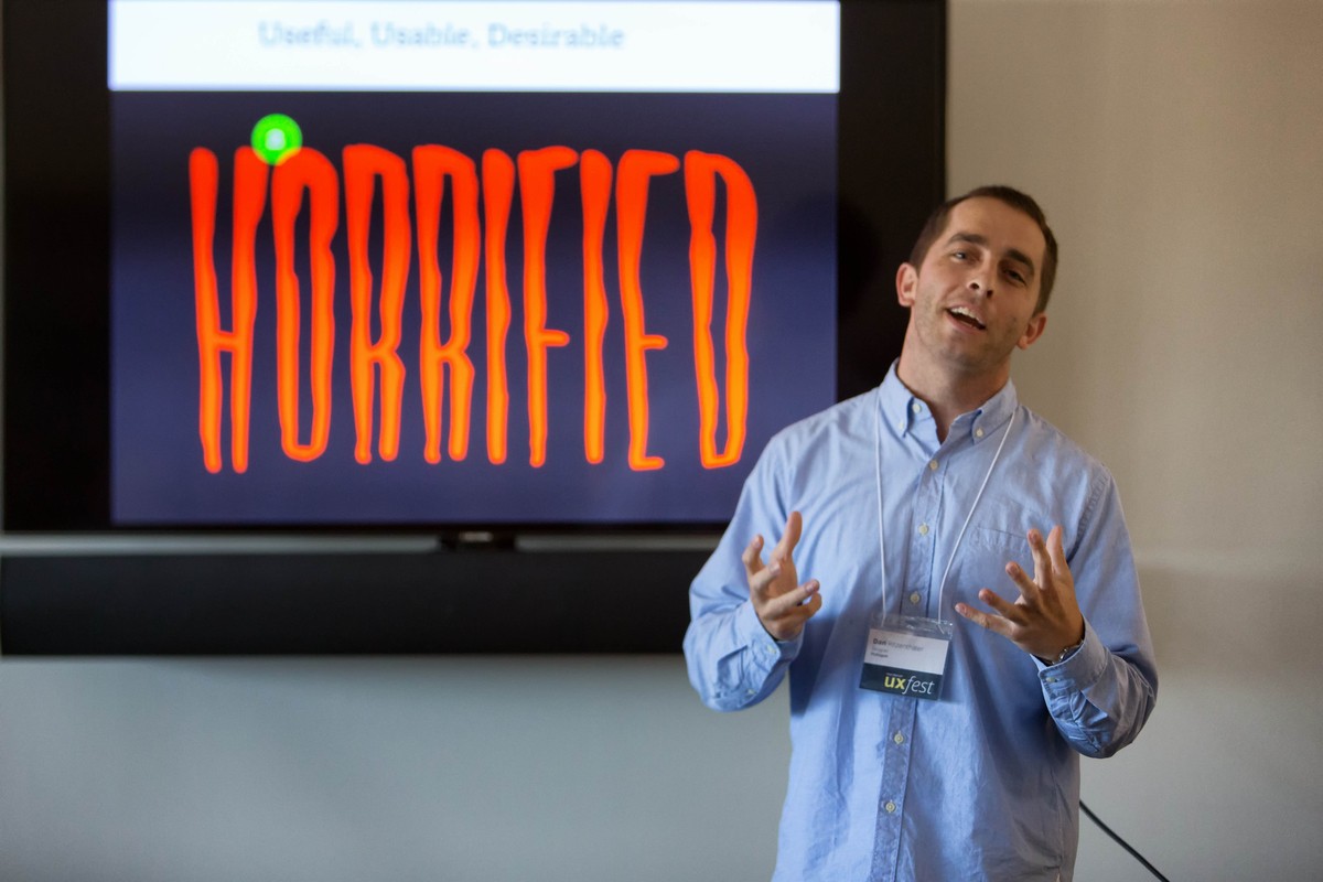An adult male speaking at an event in front of a large screen with the word 'horrified' set in a creepy font